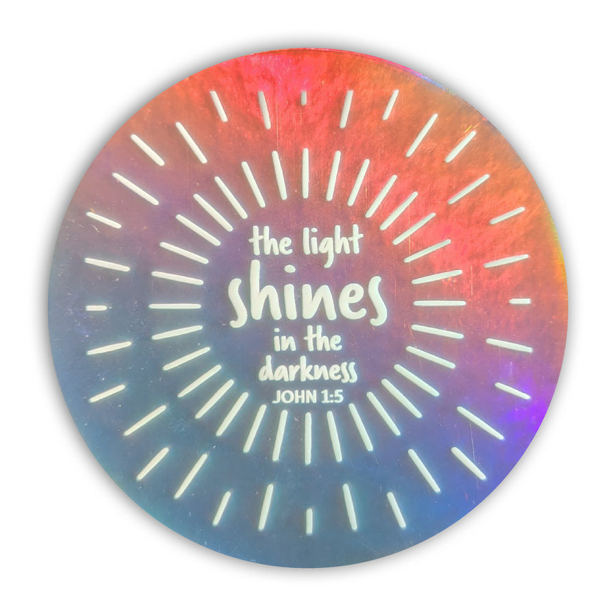 The light shines in the darkness. John 1:5. Holographic circle sticker with white Scripture design.
