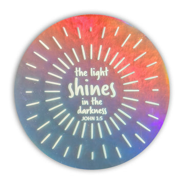 The light shines in the darkness. John 1:5. Holographic circle sticker with white Scripture design.