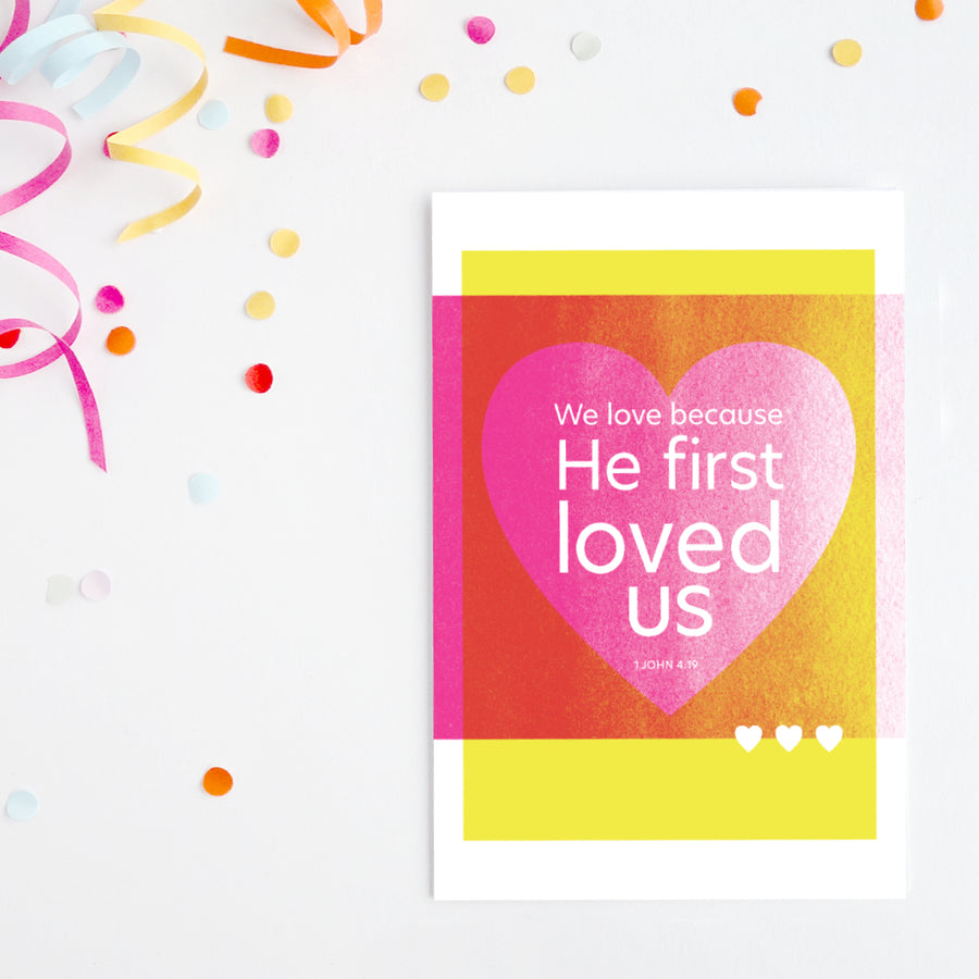 Yellow and bright pink Bible art valentine on white background with colorful confetti and ribbon. Design features a large heart and the words We love because He first loved us. 1 John 4:19.