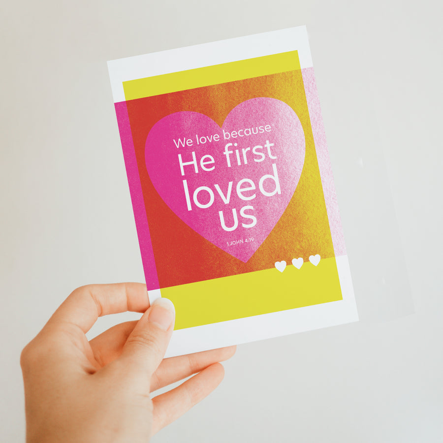 Colorful modern Scripture postcard with 1 John 4:19 We love because He first loved us. Postcard features yellow and magenta with white text.