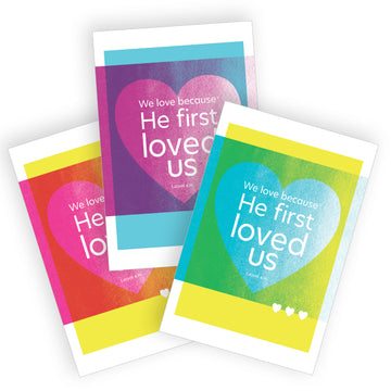 Set of Christian love postcards with 1 John 4:19 We love because He first loved us. The design features a large heart with text overlapping a solid color beneath. One postcard is magenta pink on yellow, one postcard is warm purple on sky blue, and one postcard is teal blue on yellow.