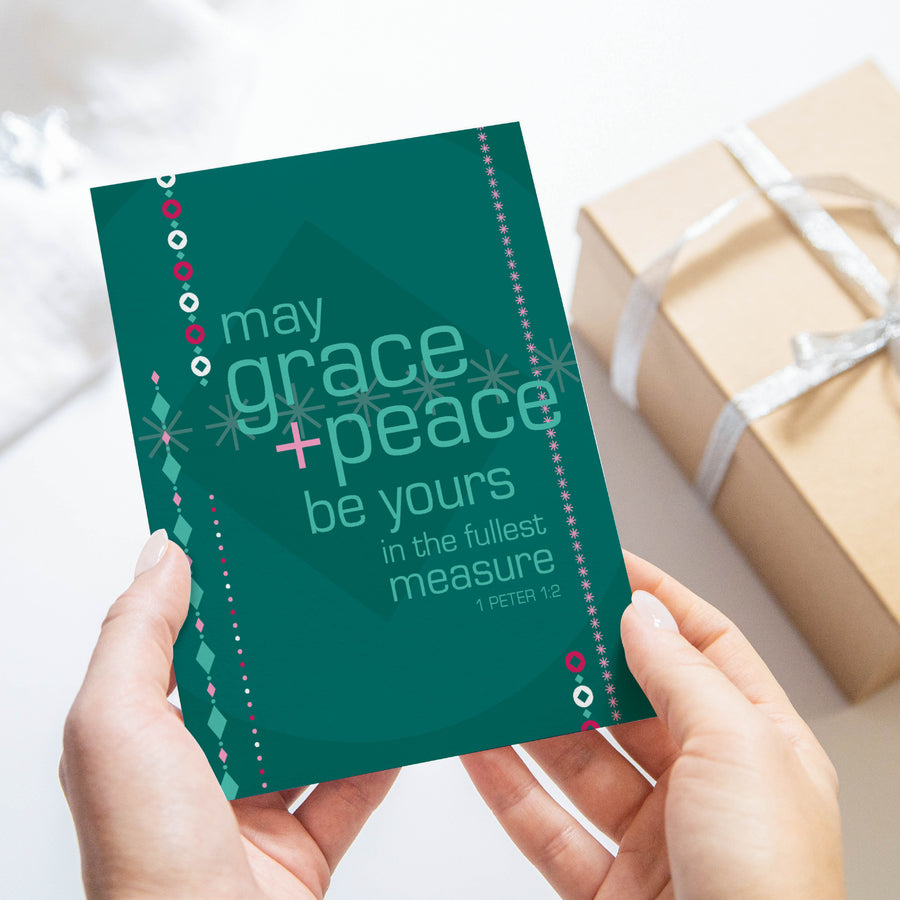 Green and pink religious Christmas card with 1 Peter 1:2 May grace and peace be yours in the fullest measure. Card is shown being held by two hands with wrapped gift in background.