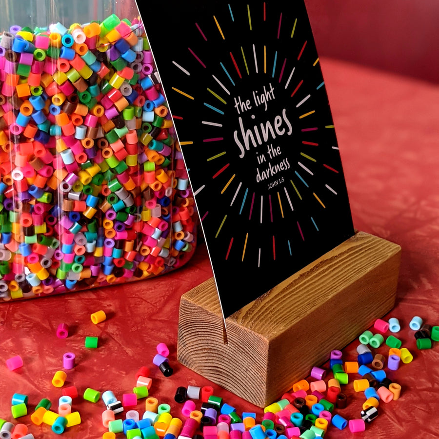 Side view of Bible verse postcard in a wood stand. Postcard is black with white text reading the light shines in the darkness. John 1:5. Tabletop is red and rainbow beads are scatterd.