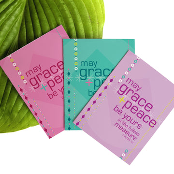 Fan of pastel Christmas cards against a large leaf. Christian Christmas cards feature text from 1 Peter 1:2 May grace and peace be yours in the fullest measure. Greeting cards are pink, seafoam green, and lavender.