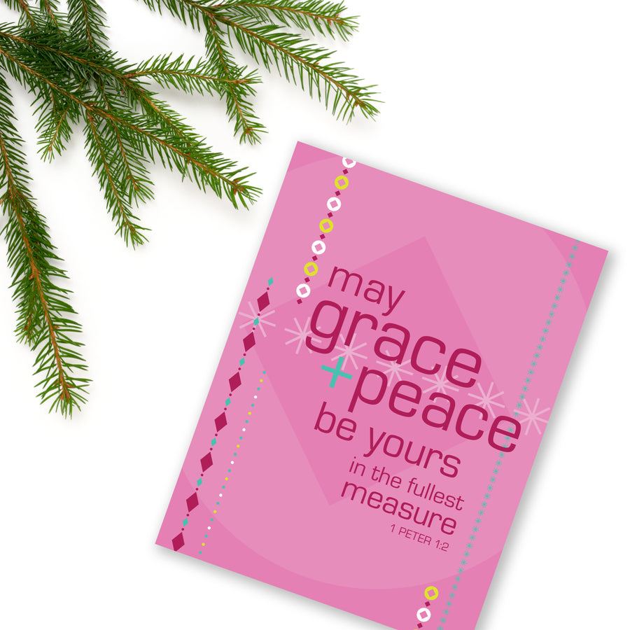 Pink Christian Christmas card angled on white background with evergreen branch in upper left corner. Bible verse greeting card features Scripture from 1 Peter 1:2 May grace and peace be yours in the fullest measure. 