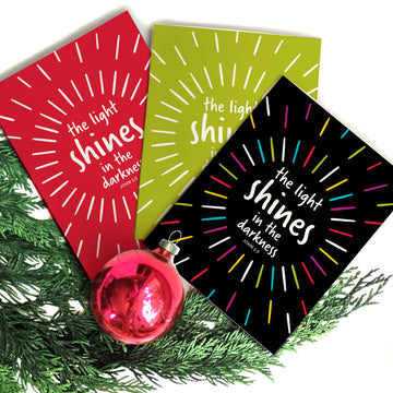 Fan of 3 Bible verse Christmas cards featuring John 1:5 the light shines in the darkness. John 1:5. Cards are red, lime green, and black with rainbow lines. Christian cards are arranged on evergreen branch with red ornament.
