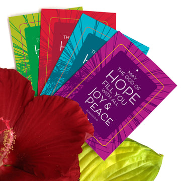Fan of 4 Christian Christmas cards arranged near a red hibiscus flower and leaf. These colorful Bible verse greeting cards feature Romans 15:13 May the God of hope fill you with all joy and peace. Cards are lime green, teal, red, and purple.