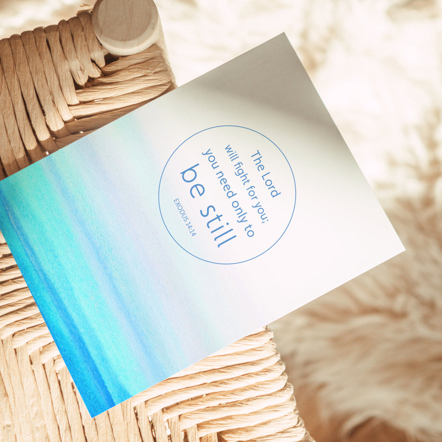 Blue and teal Scripture greeting card resting on the edge of a rattan chair. Art on the front of the card features Exodus 14:14 The Lord will fight for you; you only need to be still. Sunlight shines on the card.