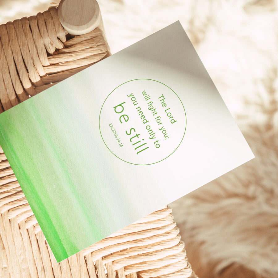 Green Bible greeting card resting on the edge of a rattan chair. Art on the front of the card features Exodus 14:14 The Lord will fight for you; you only need to be still. Sunlight shines on the card.