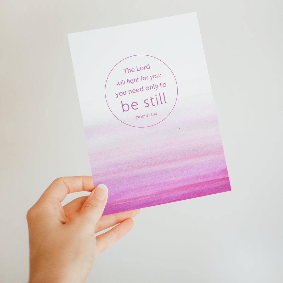 Pink Bible greeting card held up by a hand. Art on the front of the card features Exodus 14:14 The Lord will fight for you; you only need to be still. Sunlight shines on the card.