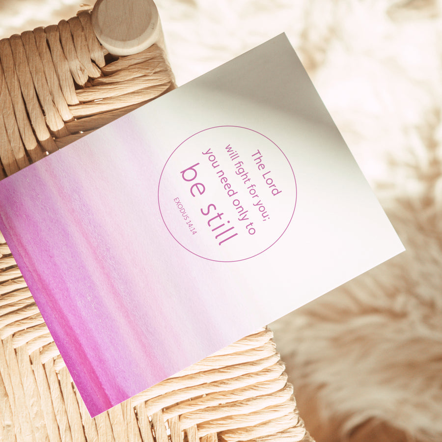 Pink Scripture greeting card resting on the edge of a rattan chair. Art on the front of the card features Exodus 14:14 The Lord will fight for you; you only need to be still. Sunlight shines on the card.
