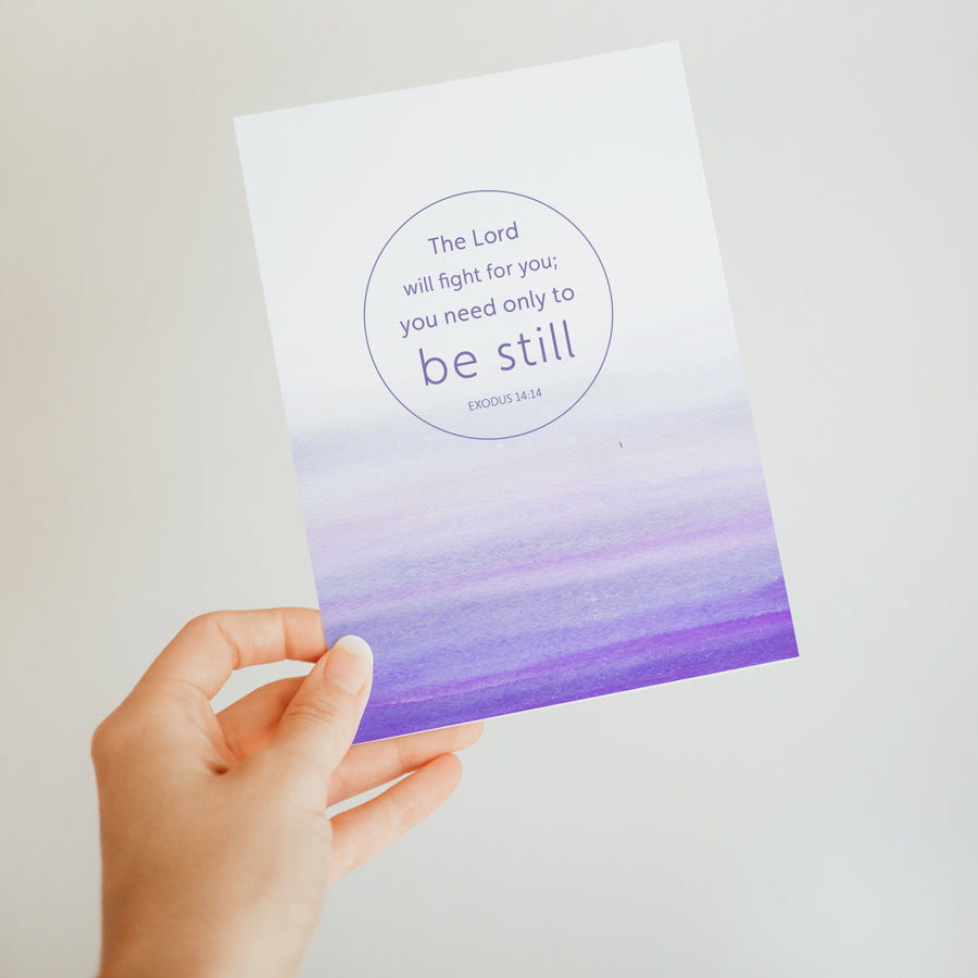 Purple Bible verse greeting card held up by a hand. Art on the front of the card features Exodus 14:14 The Lord will fight for you; you only need to be still. Sunlight shines on the card.