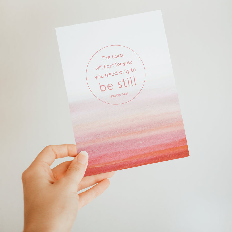 Red Christian greeting card held up by a hand. Art on the front of the card features Exodus 14:14 The Lord will fight for you; you only need to be still. Sunlight shines on the card.