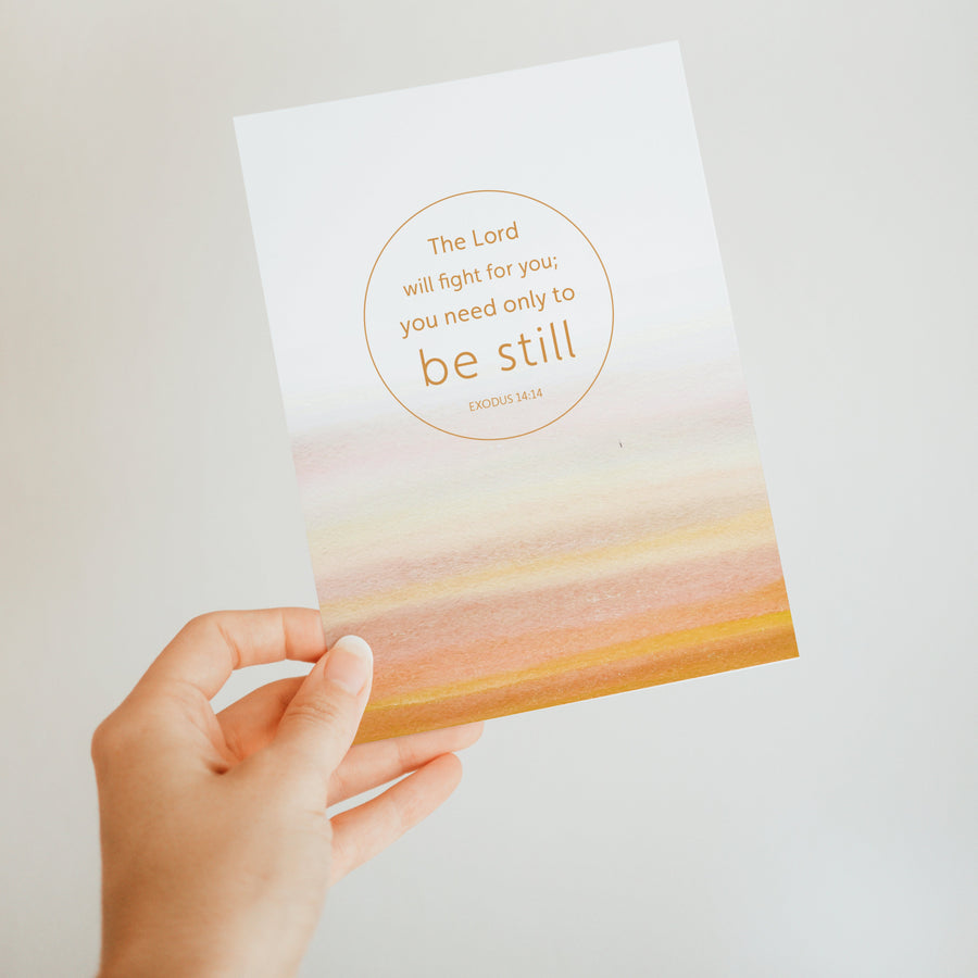 Mustard yellow Scripture greeting card held up by a hand. Art on the front of the card features Exodus 14:14 The Lord will fight for you; you only need to be still. 