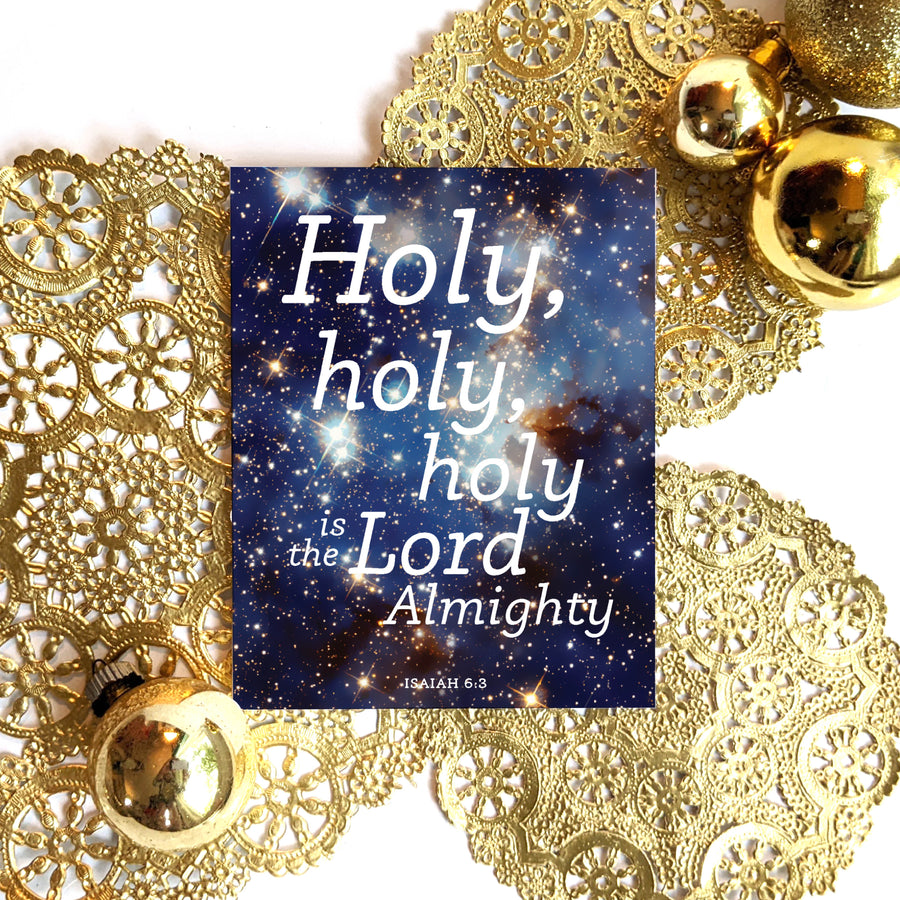 Christmas card with Bible verse resting on gold doilies with gold ornaments nearby. Card features a photo of heavens with white text on top Holy, holy, holy is the Lord Almighty. Isaiah 6:3.