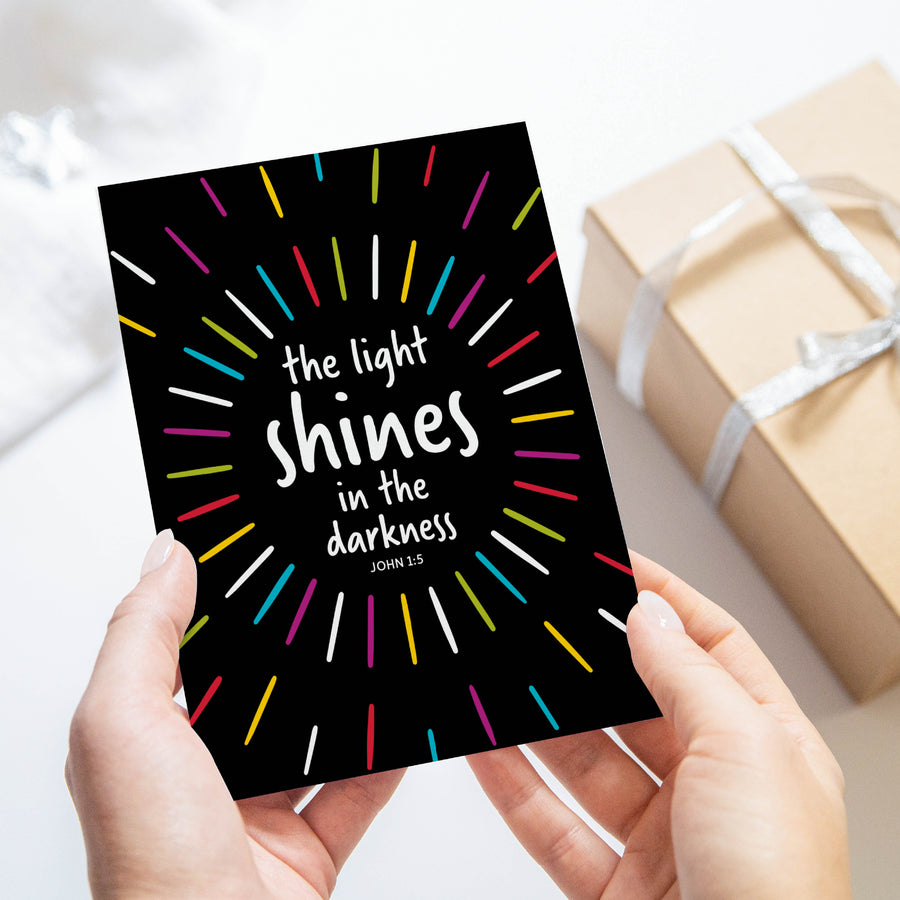 Scripture greeting card with John 1:5 the light shines in the darkness. Card shown held in hand with wrapped gifts in background.