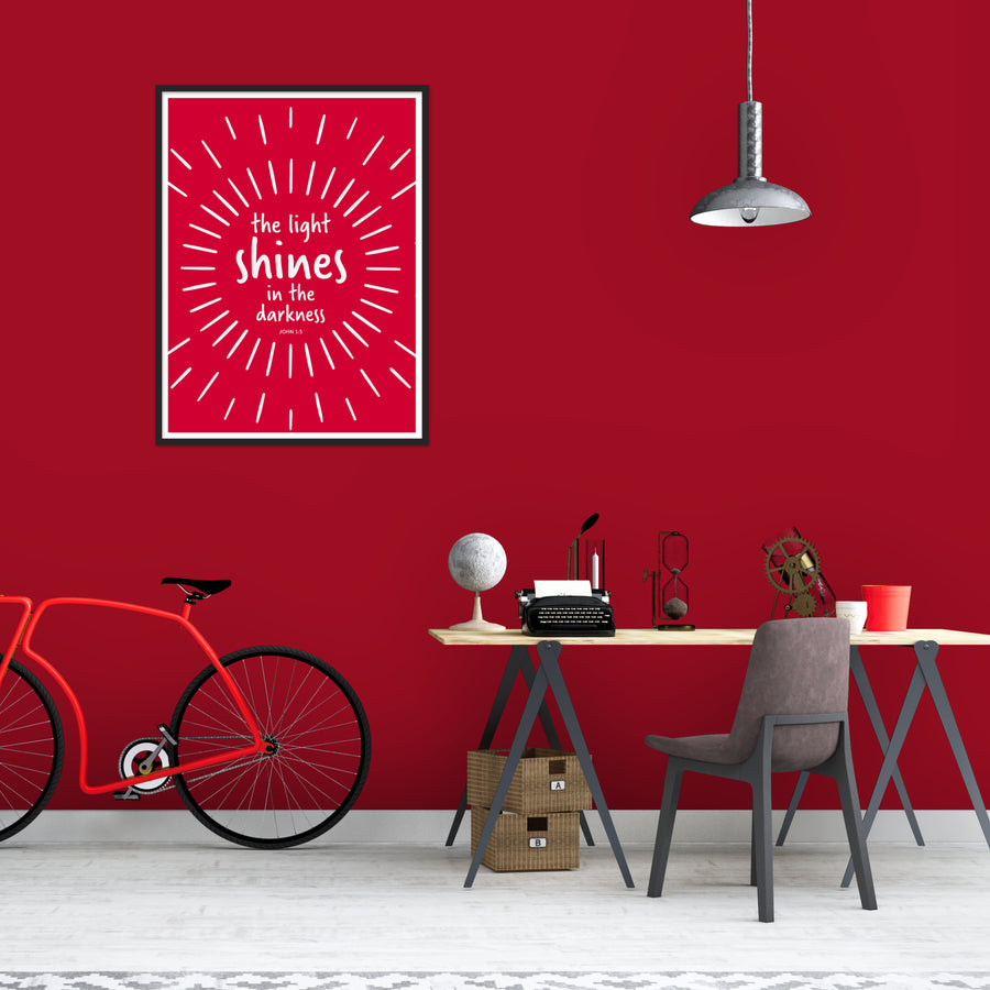 Living room with dark red walls. Bright red and white framed Christian art on wall. Art reads the light shines in the darkness. John 1:5. White lines extend outward from center circle and text. Art has white matte and is  framed in black. Modern light hangs from ceiling. Wood slab desk with metal legs. Red bike leans against wall.