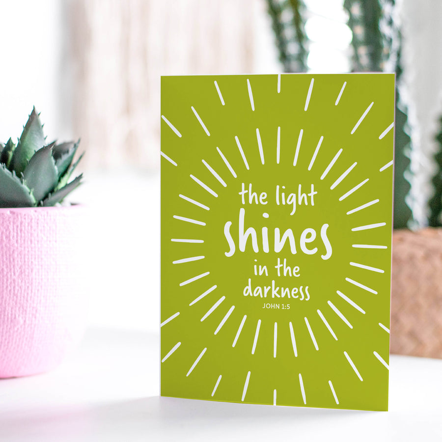 Lime green Scripture greeting card with John 1:5: the light shines in the darkness. Card shown on a tabletop next to potted plants.