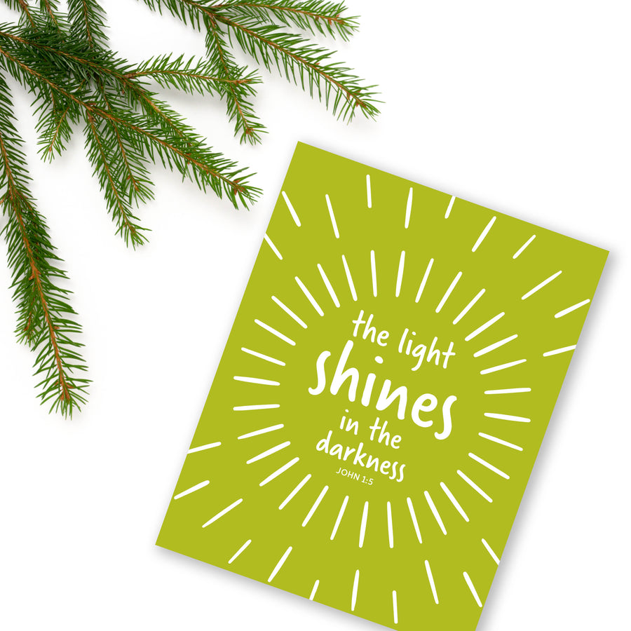 Colorful religious Christmas card with John 1:5 the light shines in the darkness. Card shown angled on white background with a pine branch in upper left corner.