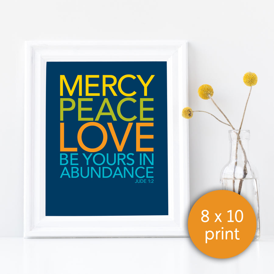 Framed Bible verse art print next to a vase with yellow flowers. Frame is white. Art is navy blue with text reading Mercy, peace, love be yours in abundance. Jude 1:2. Text in yellow, lime green, orange, and teal. Orange circle in bottom right corner labels it as 8x10 print.