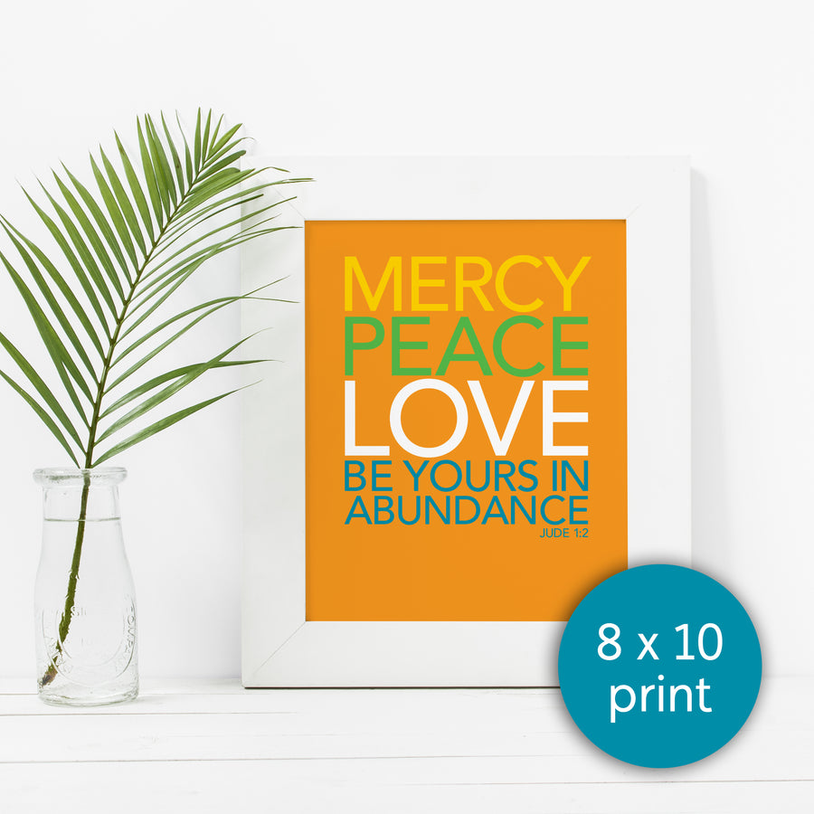 Framed Christian art print next to a vase with yellow flowers. Frame is white. Art is orange with text reading Mercy, peace, love be yours in abundance. Jude 1:2. Text in yellow, green, white, and teal. Teal circle in bottom right corner labels it as 8x10 print.