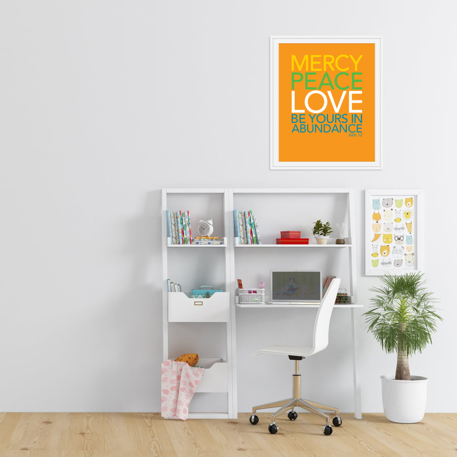 White walled room with wood floors. White leaning desk and shelves. Large artwork hanging above desk. Art is orange with text reading Mercy, peace, love be yours in abundance. Jude 1:2. Text in yellow, green, white, and teal. 