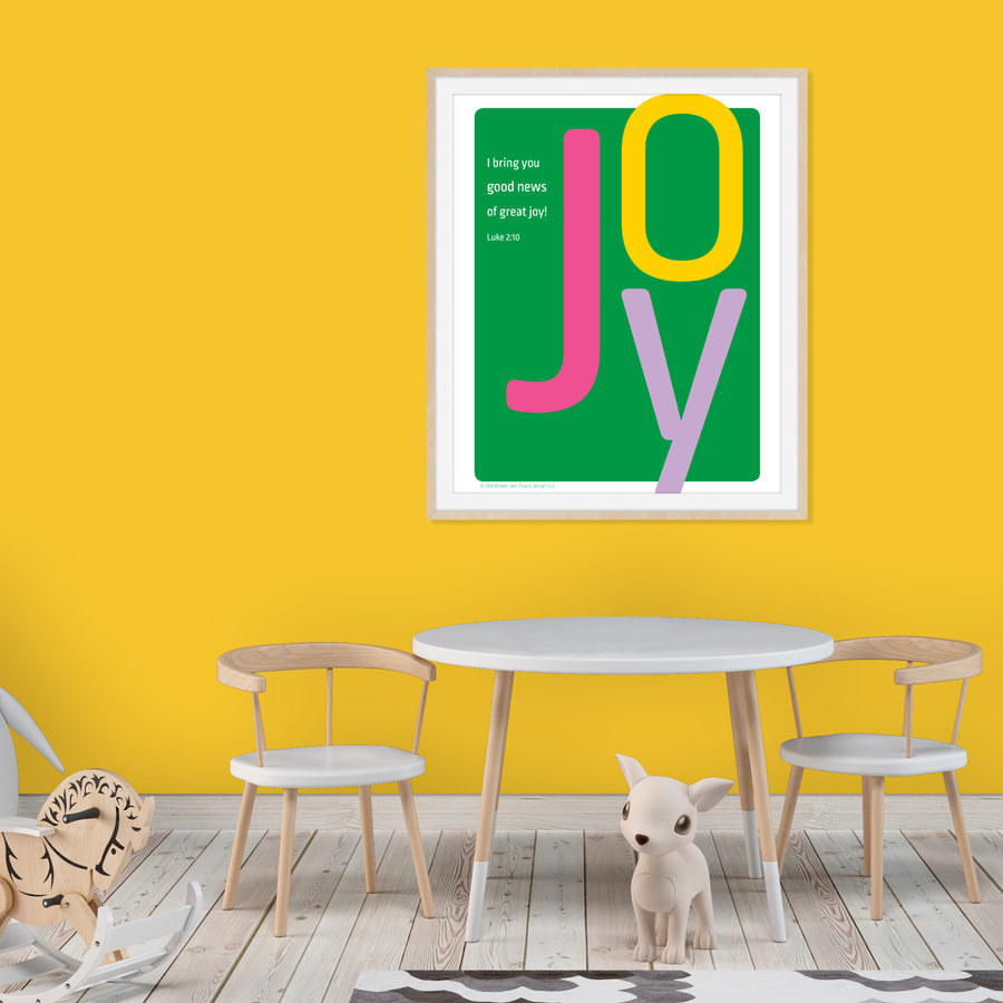 Child's room with yellow walls, wood floors, table and chairs in white with large artwork on wall. Framed kelly green Christian art features large letters spelling JOY. J is magenta. O is yellow. Y is lavender.Smaller white text reads I bring you good news of great joy! Luke 2:10. 