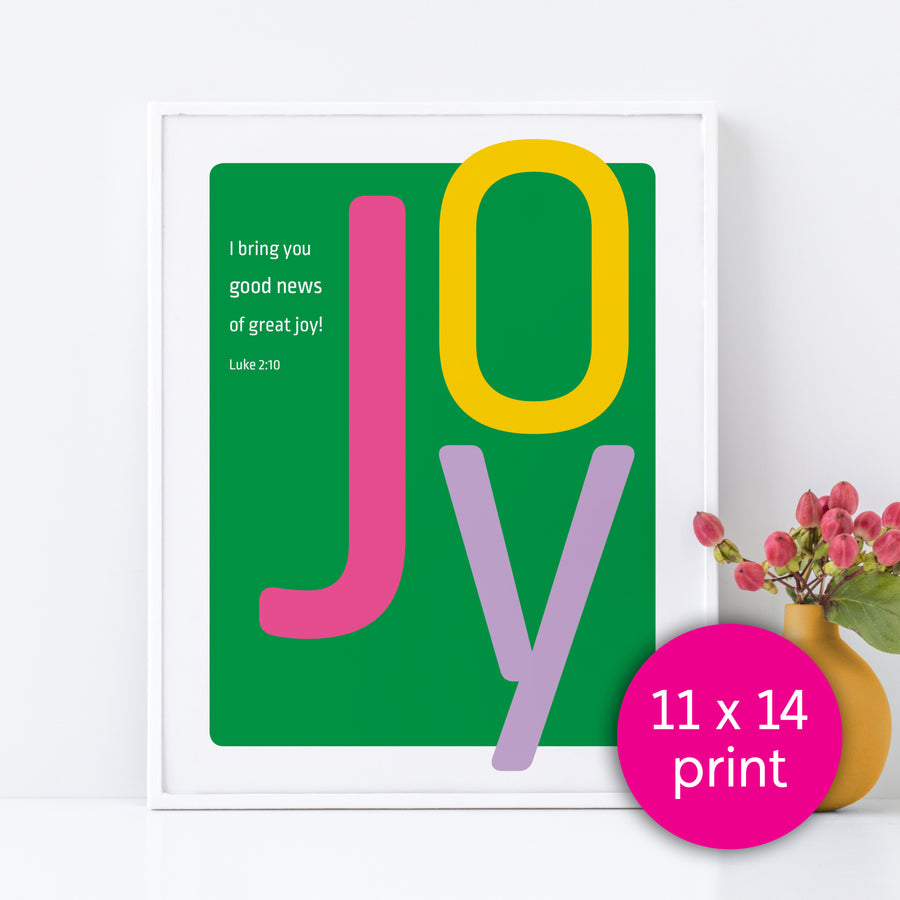 Framed Bible verse art featuring large letters spelling JOY. Smaller white text reads I bring you good news of great joy! Luke 2:10. Art in front has a kelly green background. J is magenta. O is yellow. Y is lavender. Pink circle in bottom right labels the image 11x14 print.