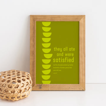Framed Bible verse art with Luke 9:17. Background is olive green. In lime green letters the type reads: They all ate and were satisfied and the disciples picked up twelve basketfuls of broken pieces that were left over. Luke 9:17. On the left side of the artwork is a stack of 12 geometric baskets extending from the top to the bottom of the art. Art is framed in a light wood frame. Small basket sits to the side.