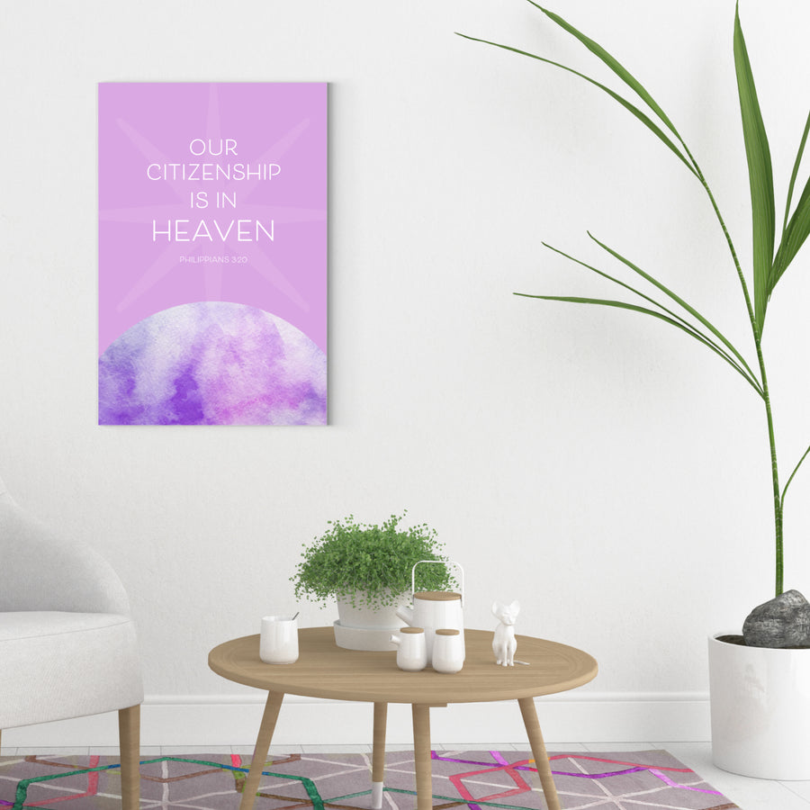 Contemporary living room scene with white walls. Large Bible verse art in lavender hangs on wall and reads Our Citizenship is in heaven. Philippians 3:20. Artwork has lavender background and white text rising above a purple and lavender watercolor planet. White chair with wood legs to the left. In the center of the image is a circular coffee table with houseplant in white pot and other white ceramic objects. Tall palm in white pot is along right edge of image.