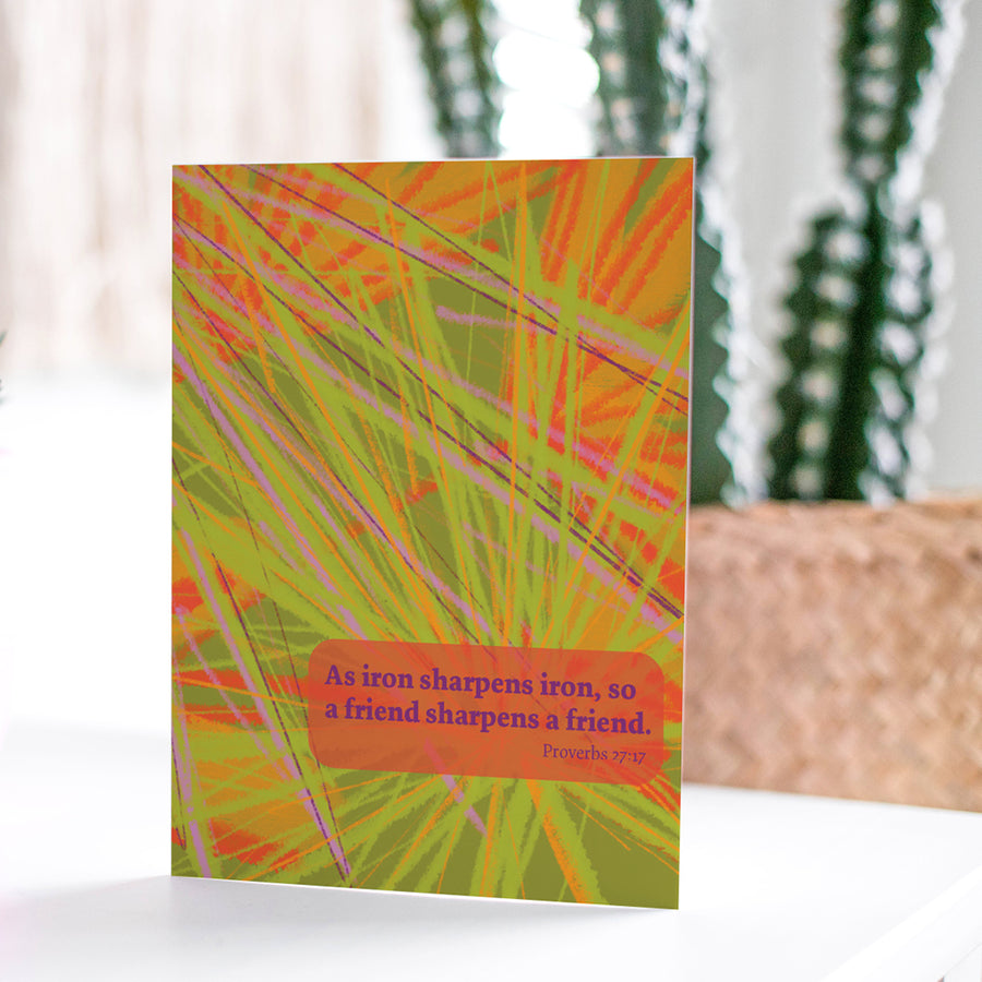 Colorful Christian greeting card with Proverbs 27:17 As iron sharpens iron, so a friend sharpens a friend. Card shown on white tabletop with cactus in the background.