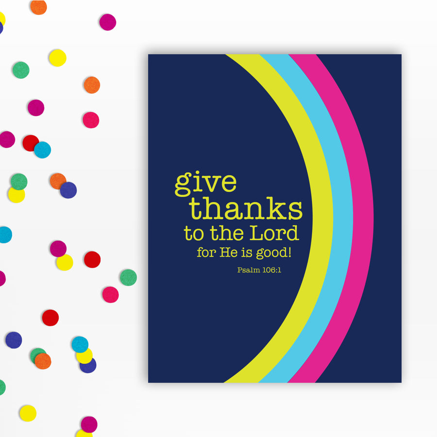 Front of a navy blue Christian greeting card with the words: give thanks to the Lord for He is good! Psalm 106:1. A curved rainbow stretches from top to bottom in magenta, sky blue, and acid yellow. Confetti to the left of the card.