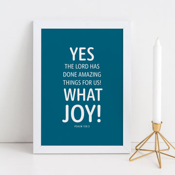 Framed Christian typography. Bible art has dark teal background with white text in rounded font reading Yes the Lord has done amazing things for us! What joy! Psalm 126:3. White candlestick in bronze holder to the right of framed image.