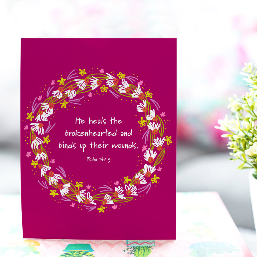 Bible verse sympathy card shown standing on a tabletop with a plant nearby. Comforting Bible verse reads He heals the brokenhearted and binds up their wounds. Psalm 147:3. Words are circled with a wreath of flowers.
