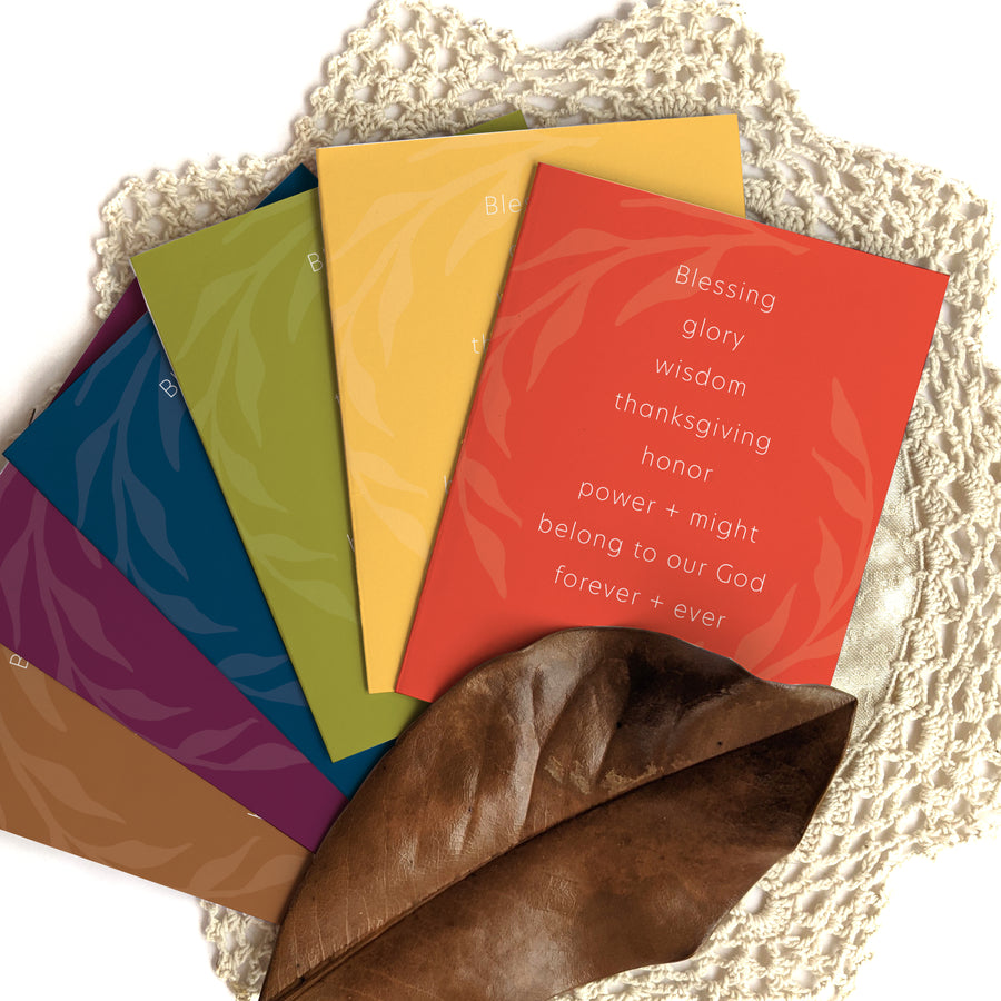 Fan of Thanksgiving greeting cards with Revelation 7:12 Bible verse. Cards are in autumn colors: brown, purple, navy blue, olive green, buttercream yellow, and orange. Cards are arranged on cream doily with a brown leaf.