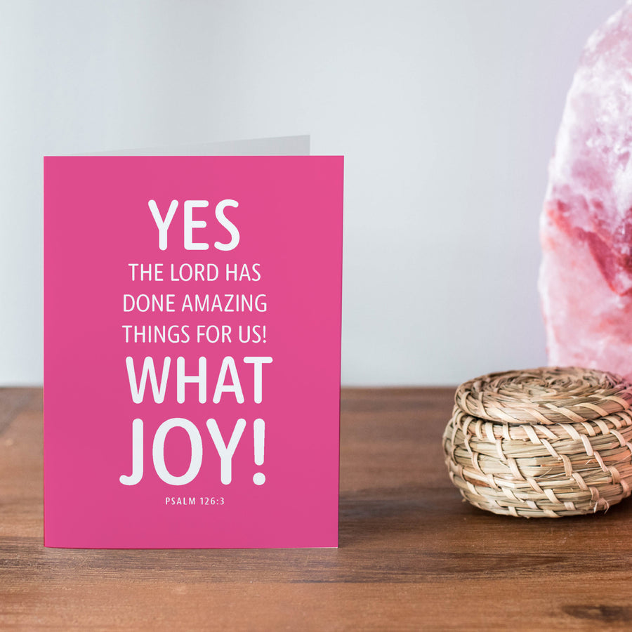 Pink and white Christian congratulations card on a wood shelf. Christian greeting card reads Yes, the Lord has done amazing things for us! What joy! Psalm 126:3.
