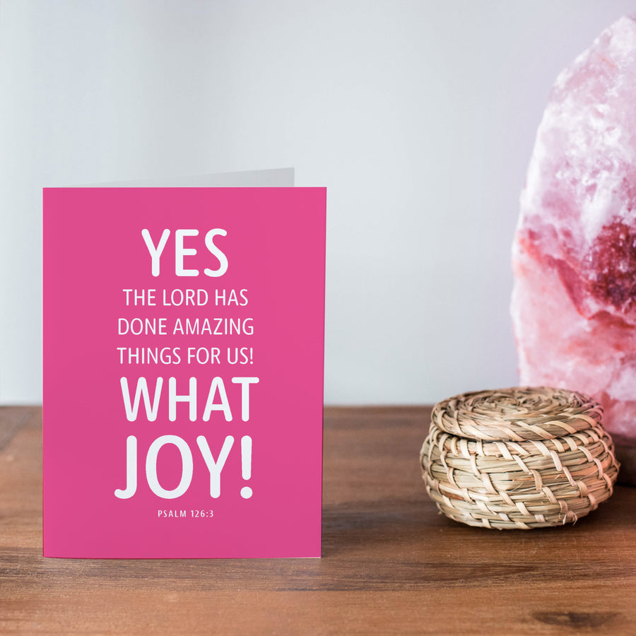 Pink and white Bible verse greeting card for celebrations. Yes, the Lord has done amazing things for us! What joy! Psalm 126:3. Card shown on a wooden shelf top near a pink rock and small basket.
