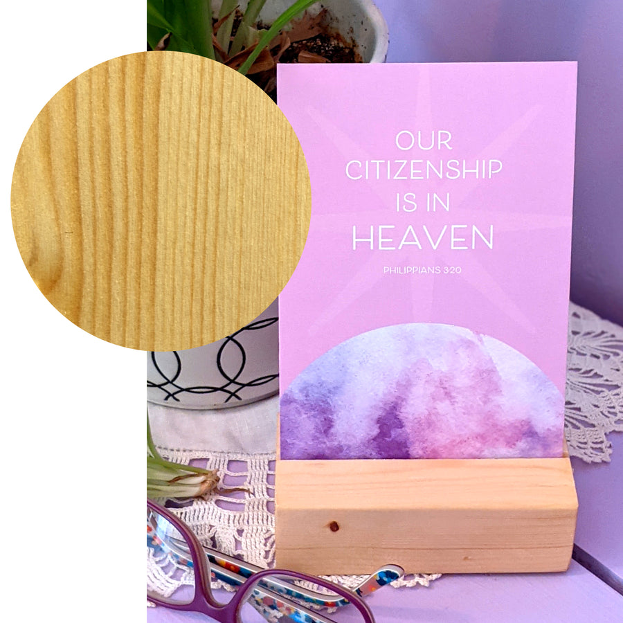 Large circle showing wood grain overlaps a photograph showing a lavender Christian postcard displayed in a wood block on a nightstand near a pair of glasses. Text on postcard reads Our citizenship is in heaven. Philippians 3:20.