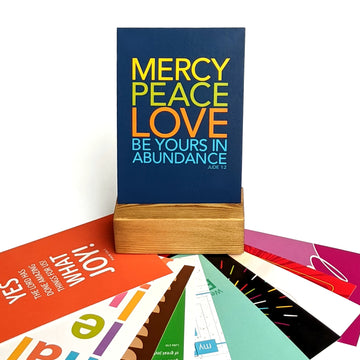 Christian postcard upright in wooden display block. Postcard is dark blue with brightly colored text from Jude 1:2 Mercy, peace, love be yours in abundance. Other postcard designs fanned out beneath it.
